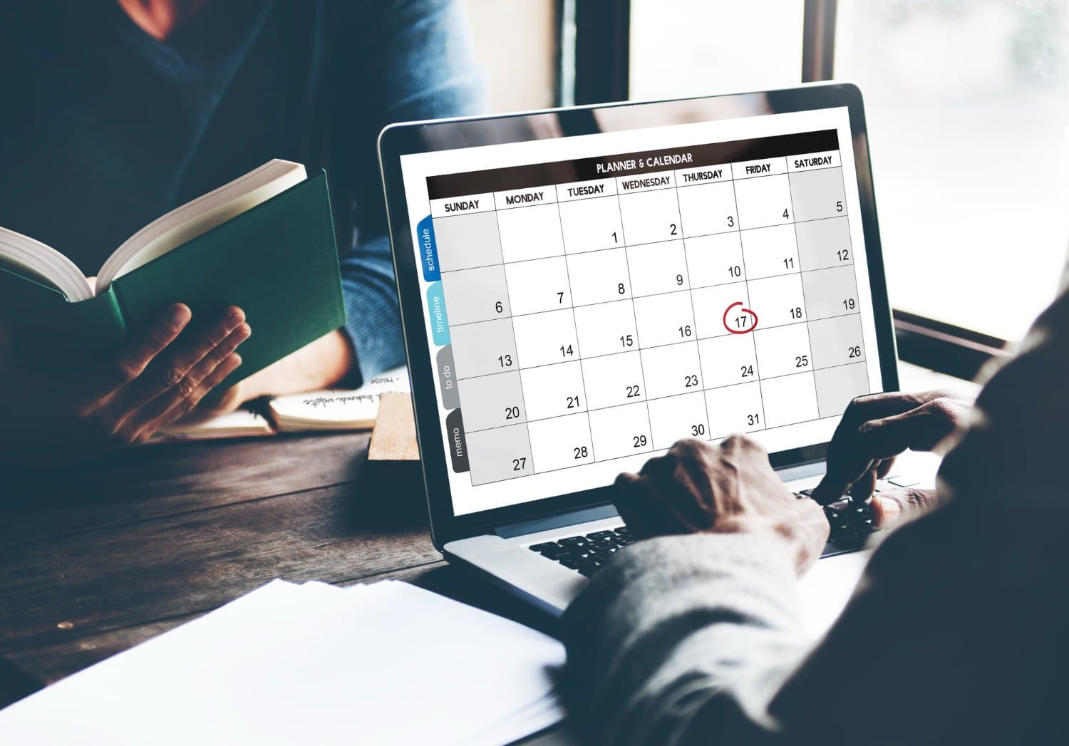 A calendar open on a laptop with a date circled, beside a person reading